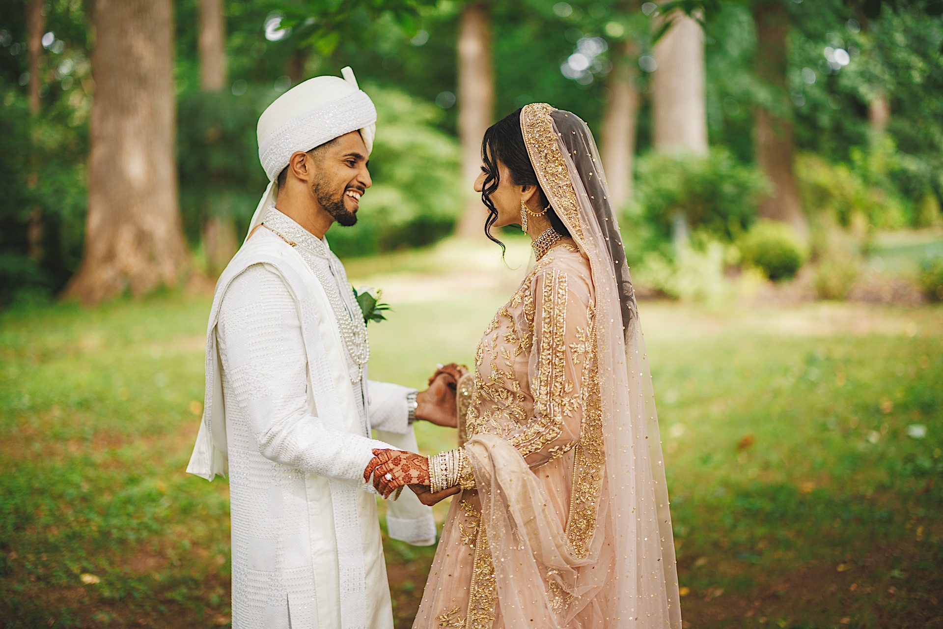 A couple dressed in traditional wedding attire smiles at each other outdoors in a park setting, surrounded by greenery, as if drafted into an auto-blooming scene of love and celebration.