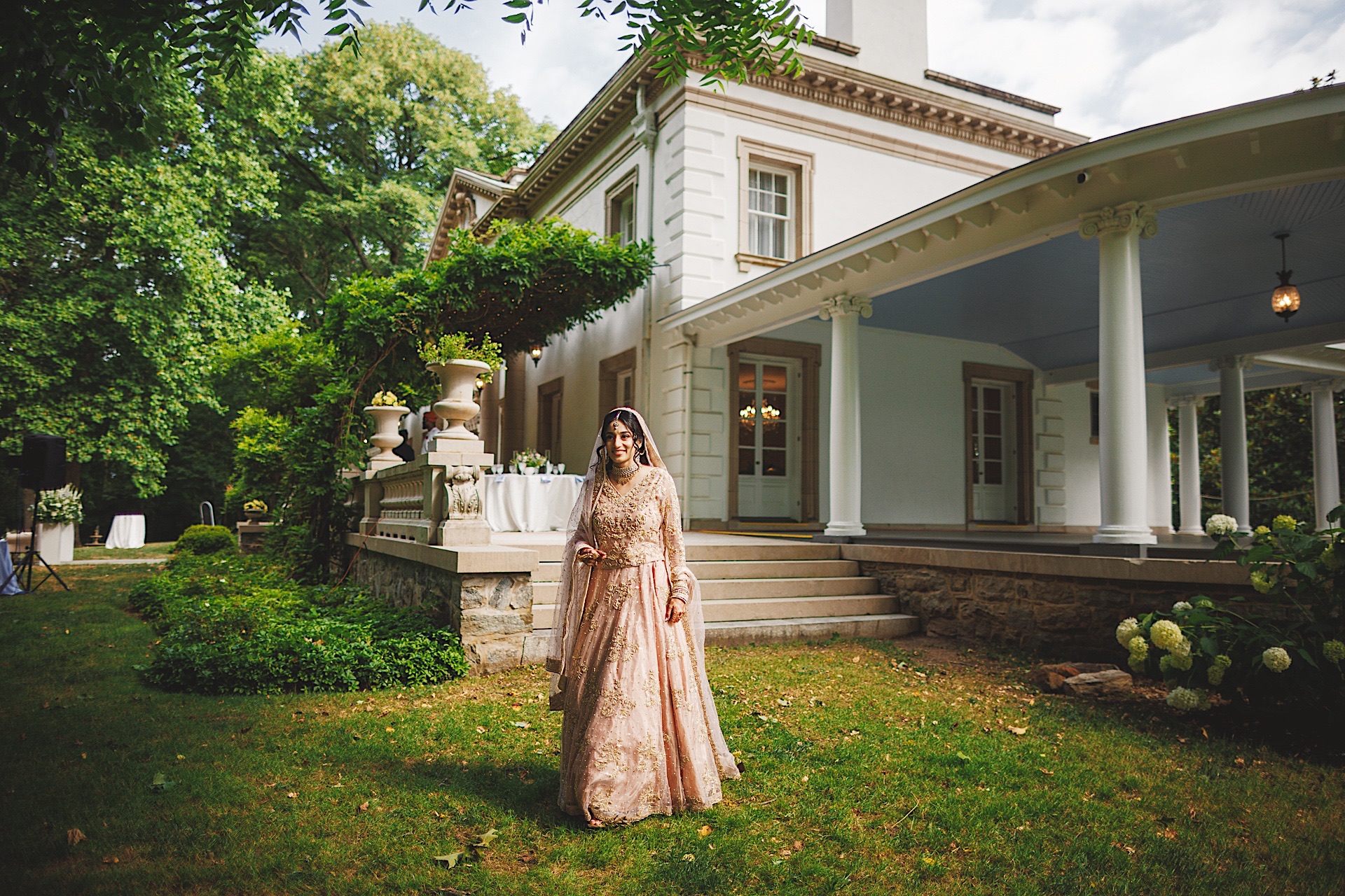 A person in a long, ornate dress stands on the lawn in front of a large, white house with columns and a covered porch, surrounded by greenery, as if they were an auto draft of an elegant era.