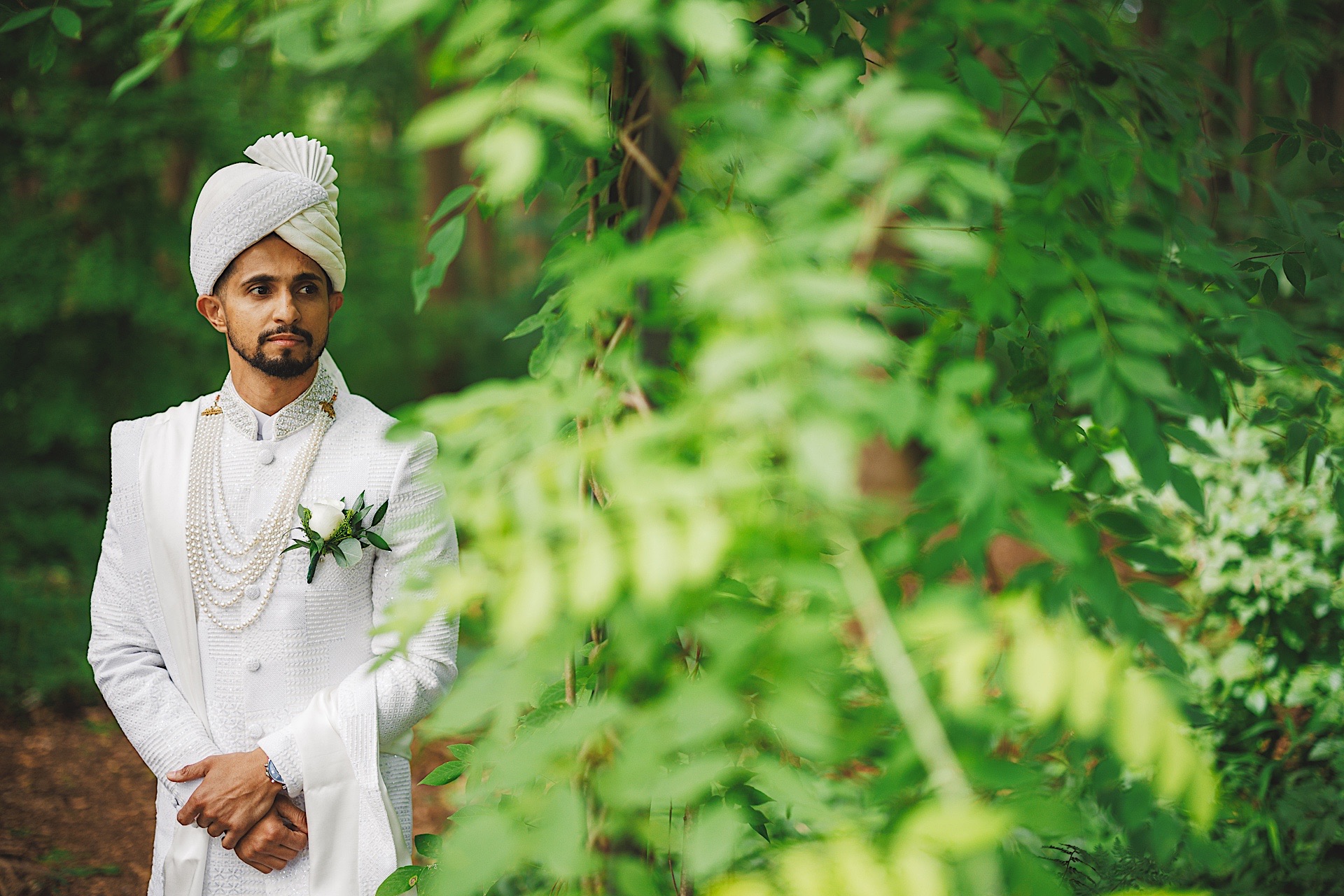 A man in a white traditional outfit and turban stands in a lush green forest, looking to the side. Sporting a string of pearls and a boutonniere on his chest, he appears almost like an auto draft of timeless elegance in this serene setting.
