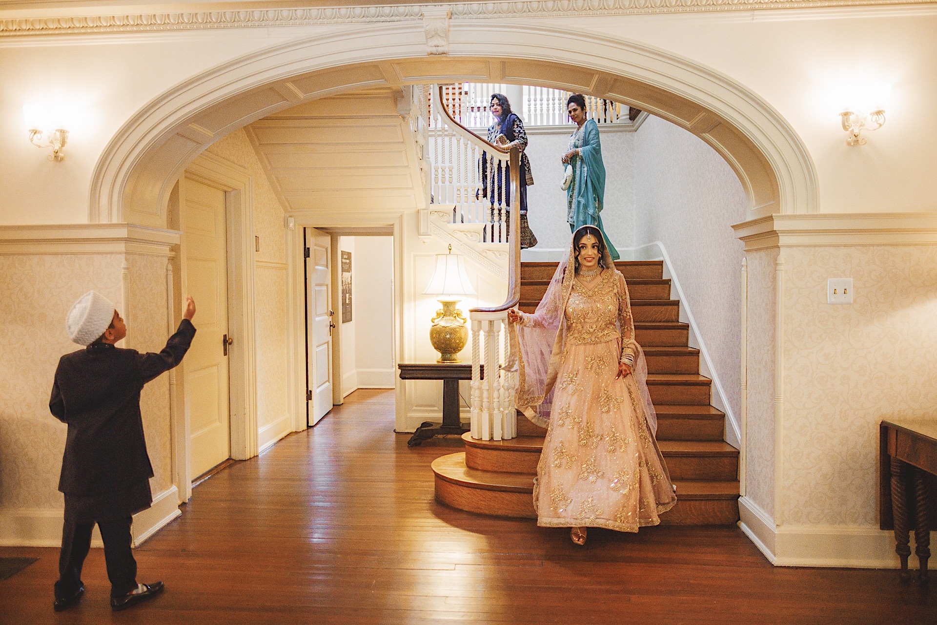 A woman in an ornate dress walks down the stairs while three people in traditional attire on the landing smile, and a young boy waves from the hallway as if auto-drafting old memories.