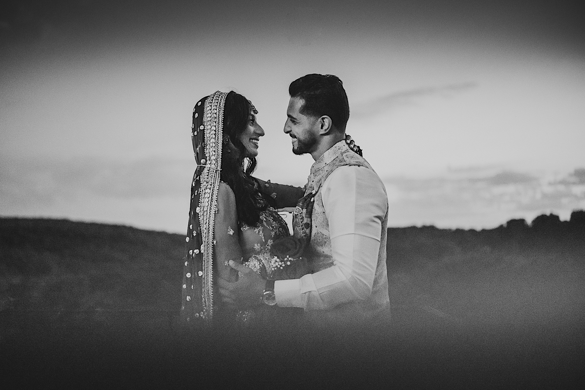 A couple dressed in traditional attire gazes at each other while embracing outdoors at dusk. The background, resembling an Auto Draft, is a blurred landscape. The image is in black and white.