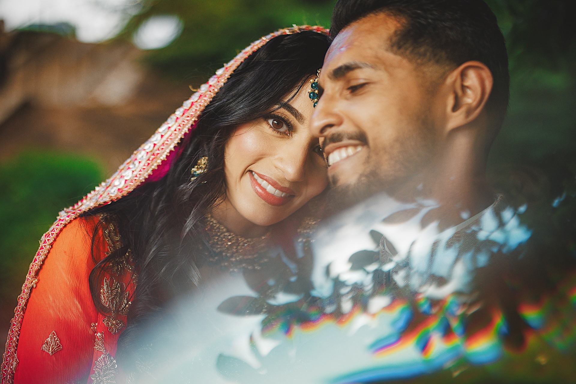 A couple dressed in traditional attire smile and lean into each other, with vibrant greenery and a rainbow-like reflection in the foreground, capturing a moment as if drafted by the magic of nature's auto draft.
