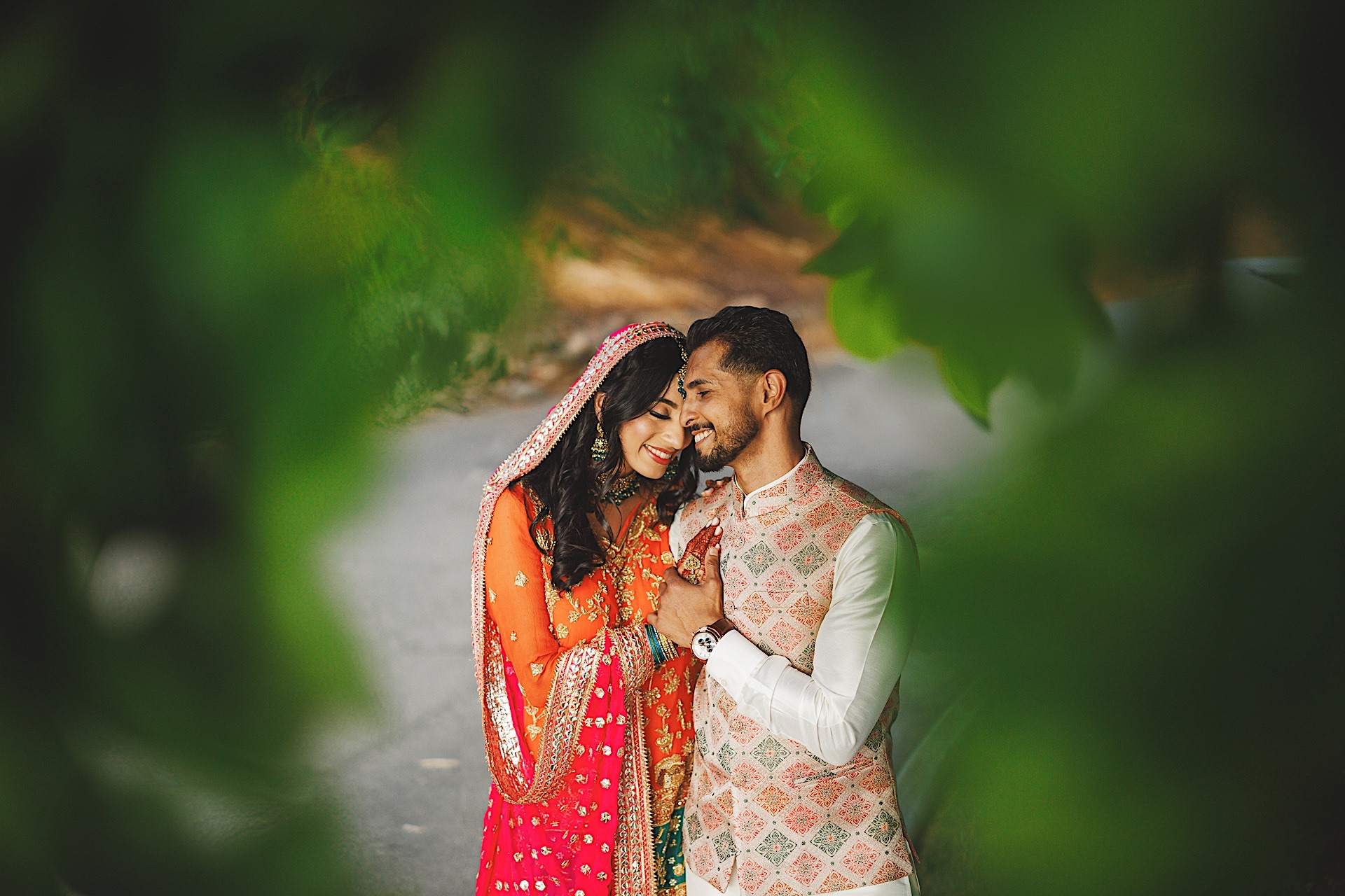 A couple, dressed in traditional South Asian attire, smiles and embraces while surrounded by greenery. The woman wears an orange and pink outfit, and the man wears a patterned vest with a white shirt, their joy captured perfectly as if on auto draft.