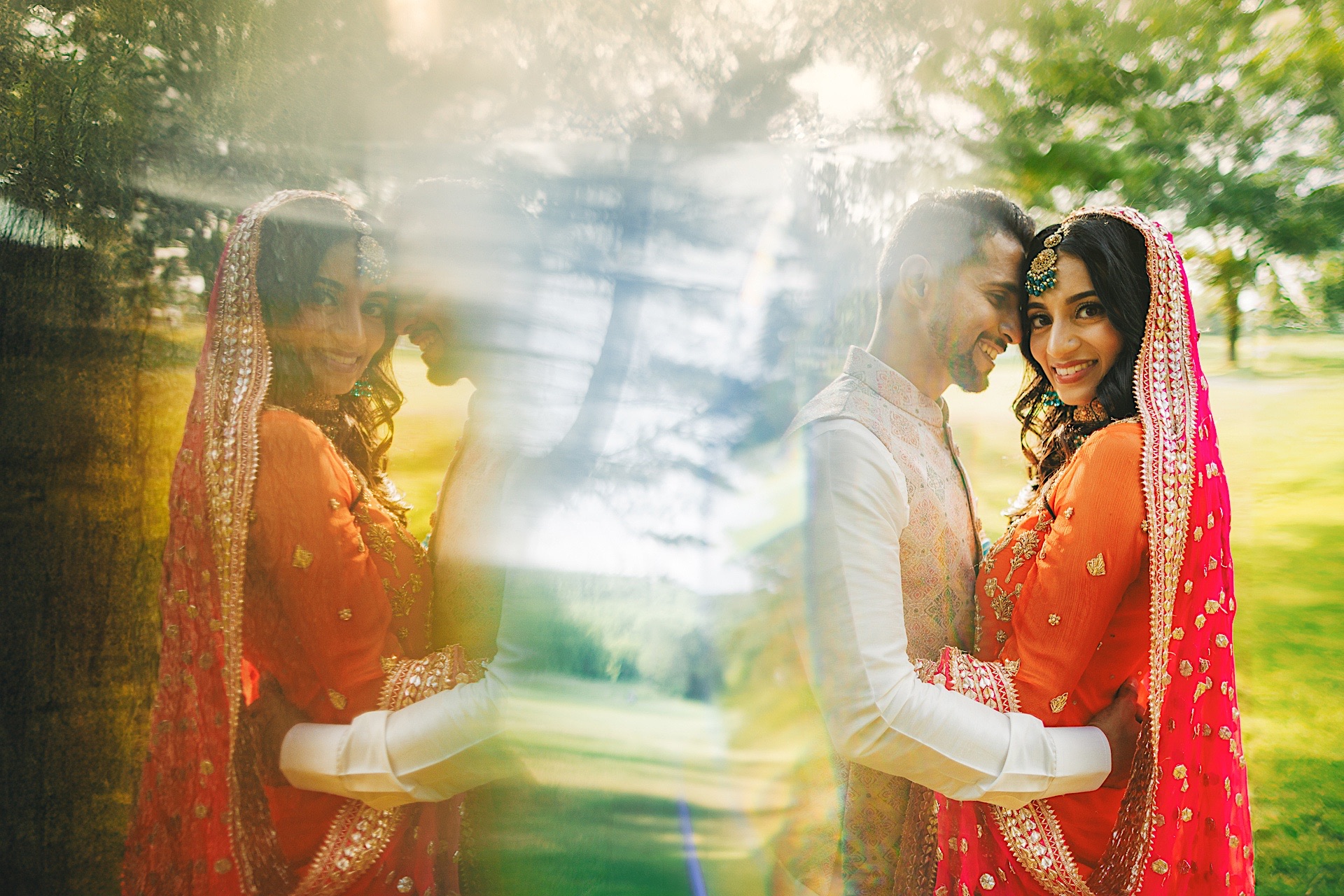 A couple in traditional clothing embrace outdoors. There's a reflection effect in the image, creating a mirrored, dreamy look. The background has a blurred, green leafy environment that feels almost like it was created with an auto draft.