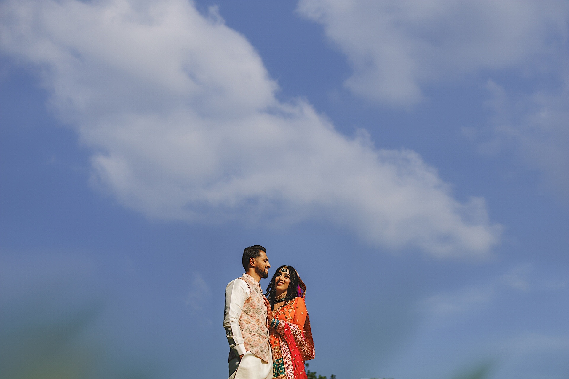 A man and woman dressed in traditional attire stand together outdoors against a backdrop of a blue sky with scattered clouds, as if part of an auto draft for a timeless portrait.