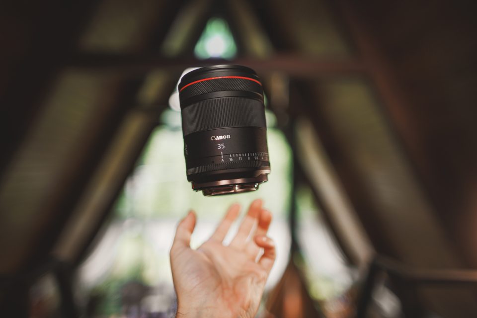 A Canon 35mm camera lens is captured in mid-air above an outstretched hand inside a rustic, wood-paneled structure, seemingly as if suspended by auto-draft magic.