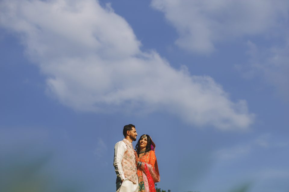 A couple stands closely together, smiling under a bright blue sky with scattered white clouds. The man wears a light-colored outfit, and the woman is dressed in a vibrant, traditional garment.