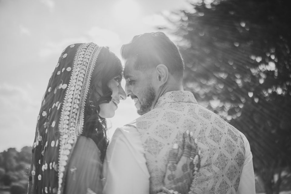 A black and white photo of a couple facing each other, the woman in a decorated bridal veil and the man in a patterned vest, with trees and sunlight in the background.