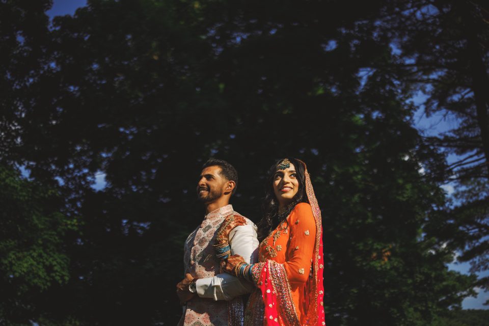 A couple stands side by side outdoors, wearing traditional South Asian attire. The man is in light-colored clothing, while the woman is in a vibrant orange outfit with intricate decorations. Trees are in the background.