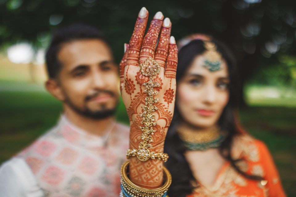 A woman with intricate henna designs on her hand adorned with jewelry, holds it up in focus, while a man stands blurred in the background.