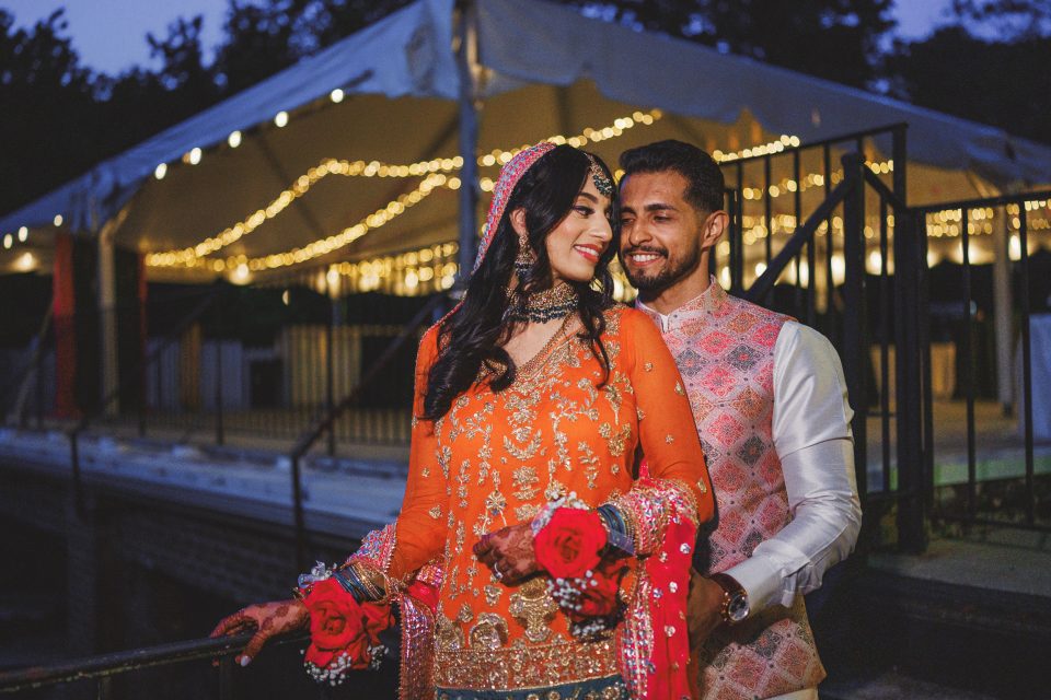 A couple dressed in vibrant traditional attire poses together under string lights at an outdoor venue in the evening. They are smiling and holding red roses.