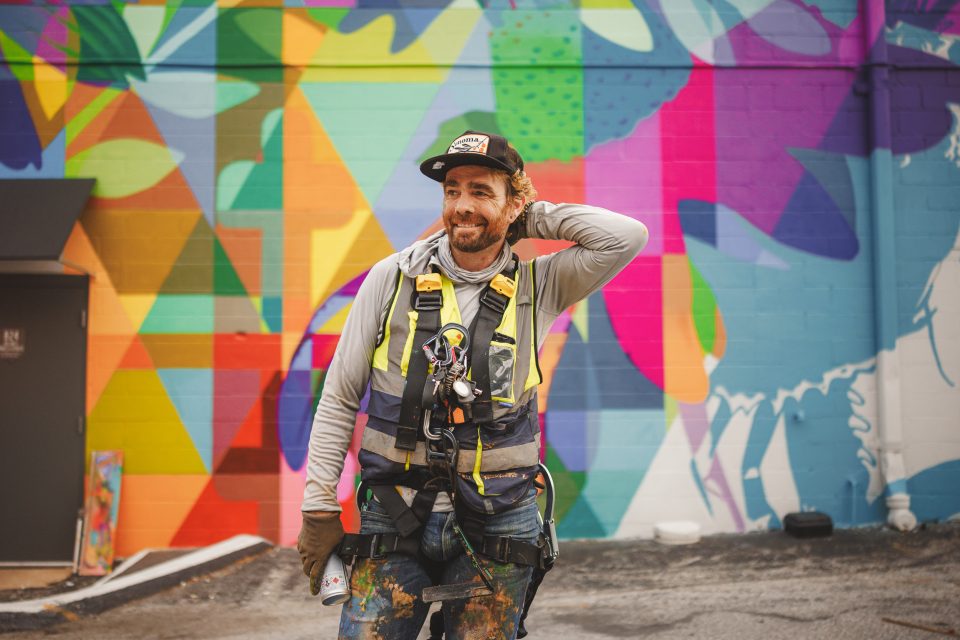 A man wearing a safety vest and paint-stained clothing stands in front of a colorful, abstract mural, holding a spray paint can.