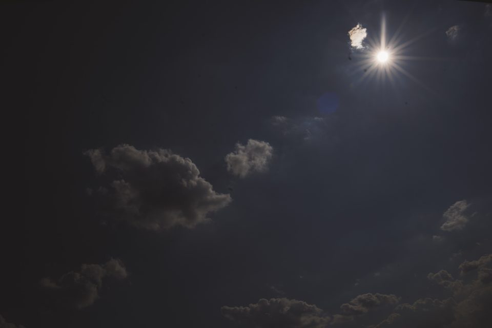 The image shows a dark sky with scattered clouds and the sun partially obscured by one cloud, creating a bright glare.