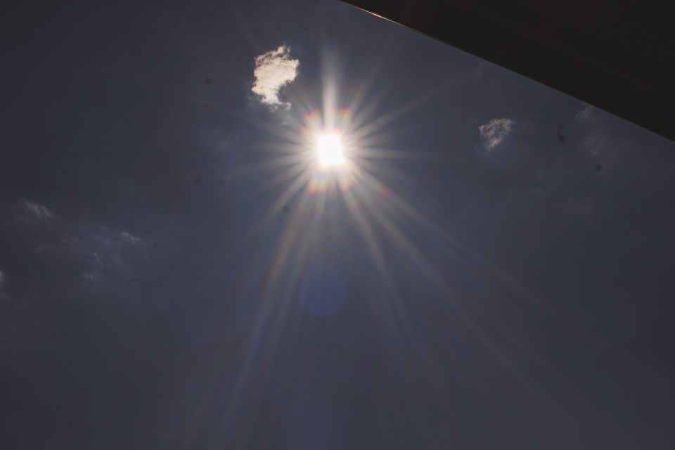 Bright sun with rays extending outward is partially covered by a small cloud in a dark sky with scattered clouds.