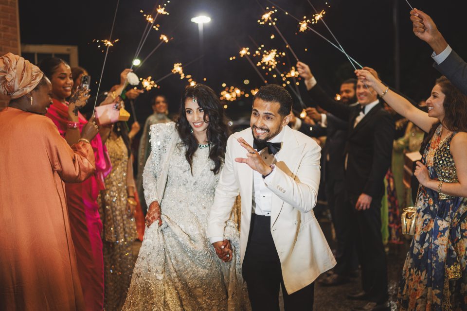 A couple, dressed in wedding attire, walks hand-in-hand under a canopy of sparklers held by friends and family in celebratory attire during a nighttime wedding event, perfectly captured through the crystal-clear optics of the Canon 35 1.4 VCM lens.