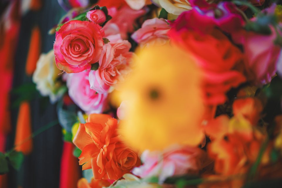 A close-up of a colorful floral arrangement featuring pink and orange roses, with a prominent unfocused yellow flower in the foreground.
