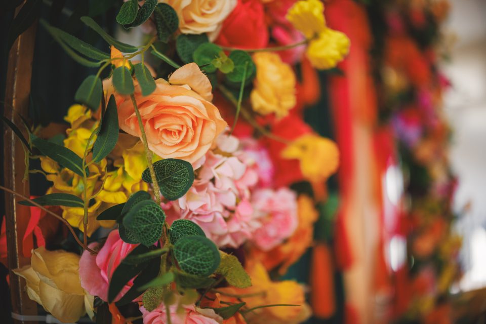 Close-up of a colorful flower arrangement featuring roses in shades of orange, pink, and yellow, surrounded by green leaves and other vibrant blooms.