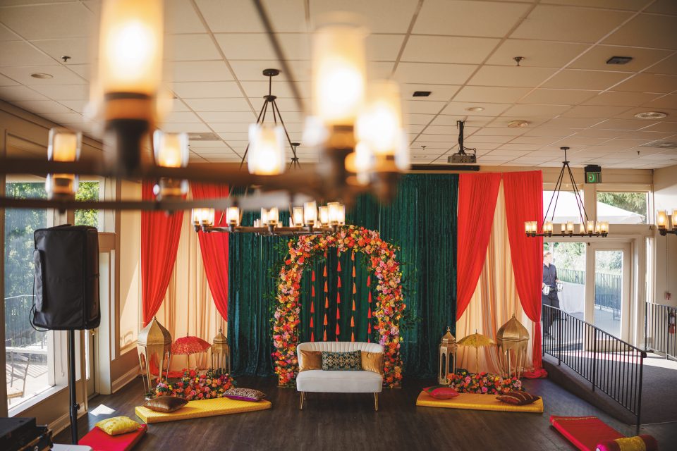 A decorated stage features a white couch in front of a vibrant backdrop with red, green, and gold drapery, flanked by colorful flowers. Floor seating and lanterns are arranged around the stage.