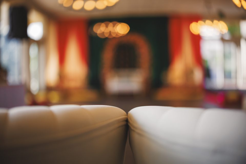 Blurred image of a decorative indoor event space with soft lighting, featuring a white tufted seat in the foreground and colorful draped fabric in the background.