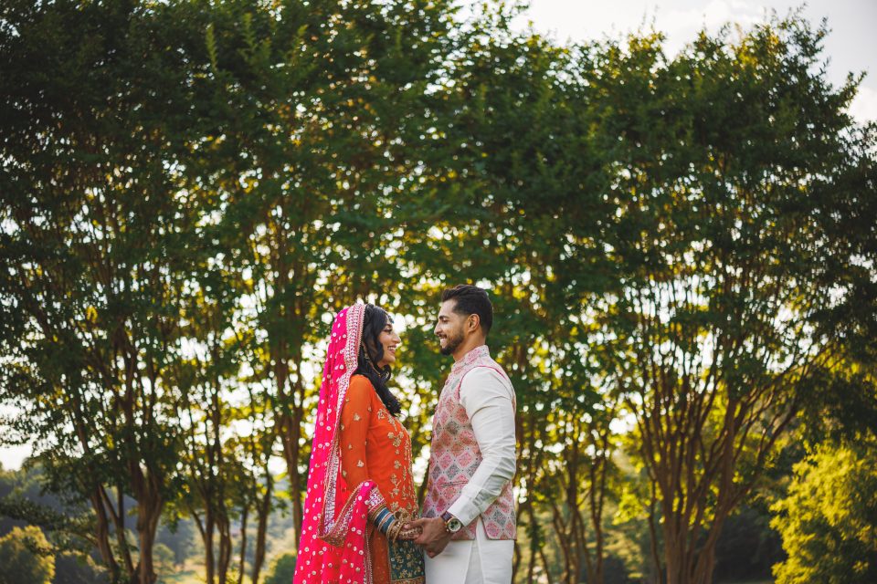 A couple stands facing each other and holding hands outdoors in front of tall green trees. The woman is wearing a pink traditional outfit and the man is dressed in white with a patterned vest.