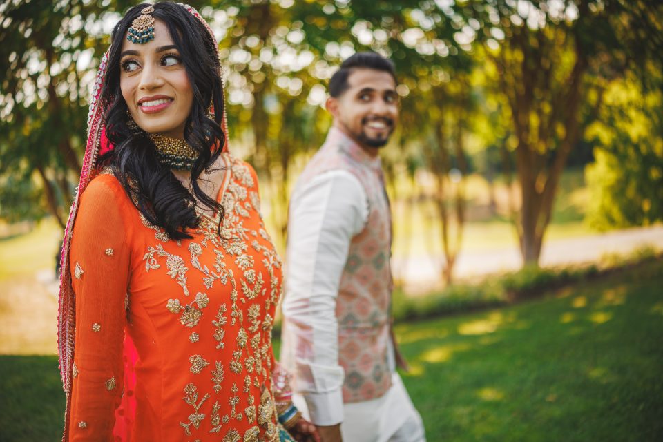 A woman in an orange embroidered dress and a man in light traditional attire hold hands and walk outdoors on a sunny day, with trees in the background.