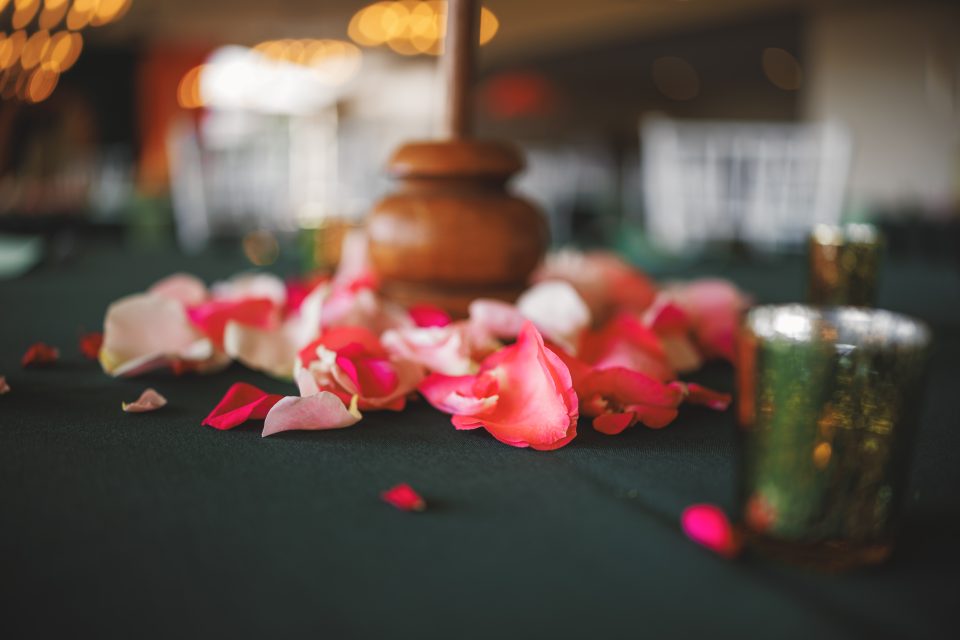 Close-up of rose petals scattered on a dark green tablecloth with candles and a wooden decoration in the background.