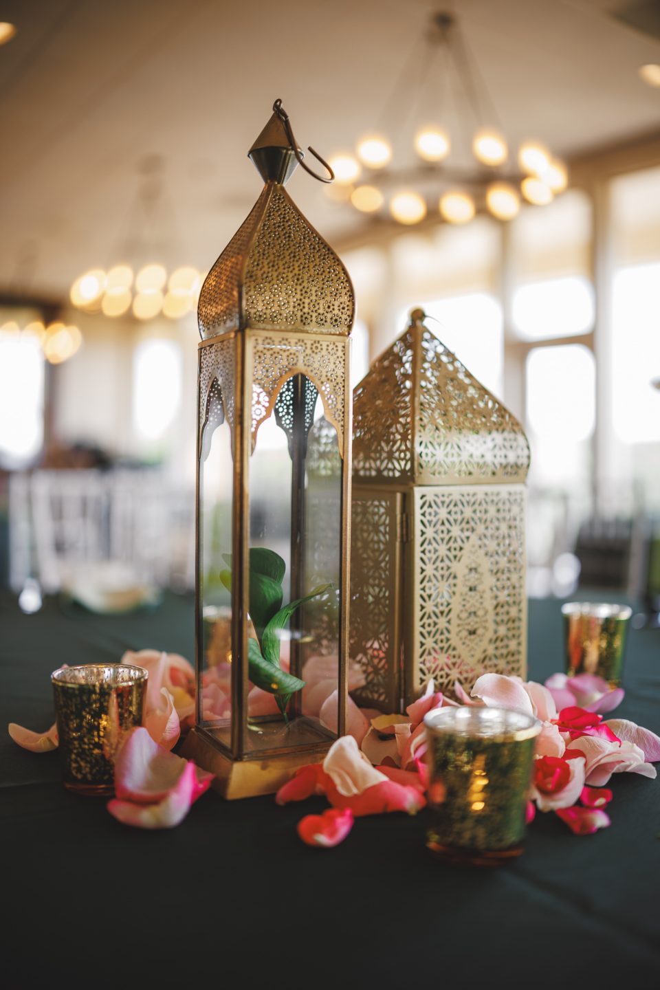 Two ornate golden lanterns with intricate patterns are placed on a table, surrounded by scattered pink and white flower petals and small votive candles.