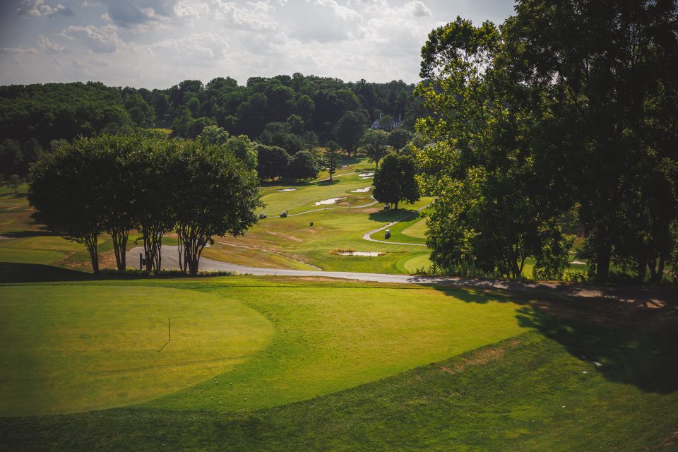 A golf course with well-maintained green fields, trees, and a winding path is seen under a partly cloudy sky.