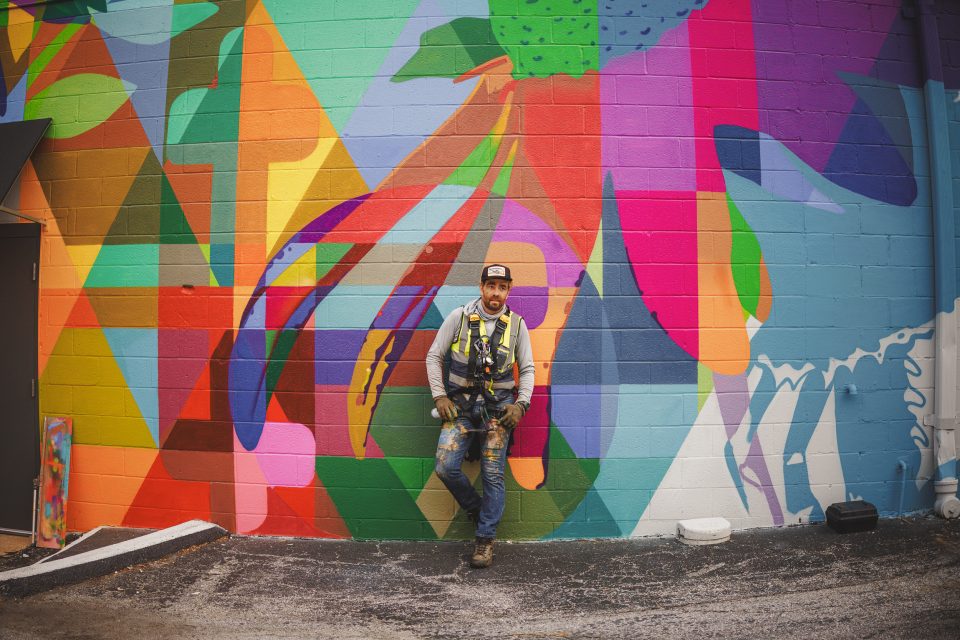 A man wearing a cap and harness leans against a brightly colored geometric mural on a building wall, holding a spray can, appearing to be the mural’s painter. Captured through the precision of a Canon 35 1.4 VCM lens, the scene blends artistry with vivid detail.