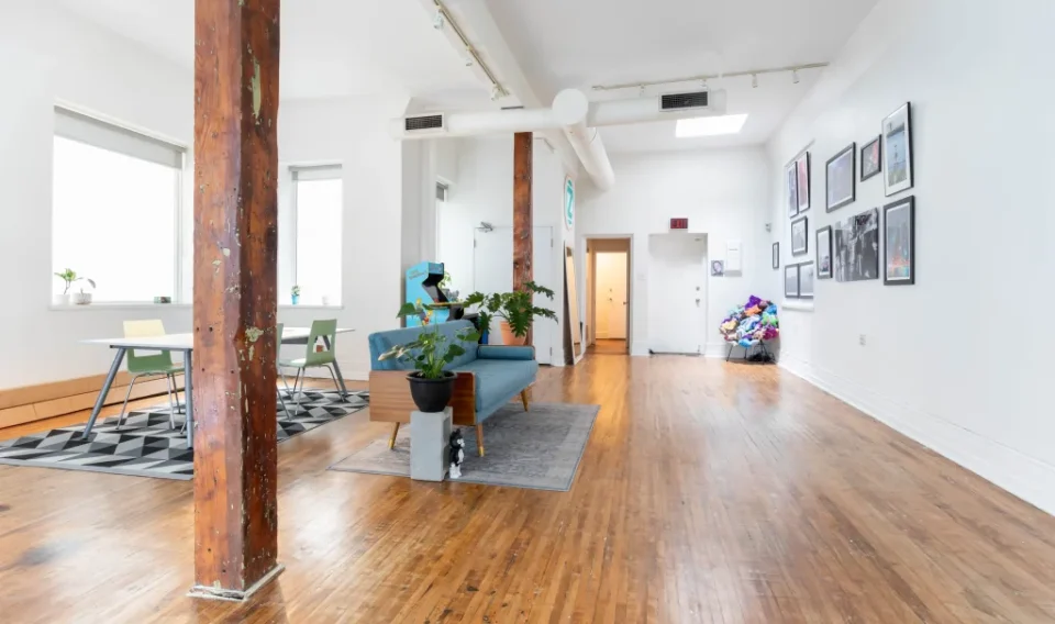 A spacious room with wooden floors, white walls, and exposed wooden beams. It features a blue sofa, potted plants, a table with chairs, framed pictures, and a pile of colorful plush toys in the corner.