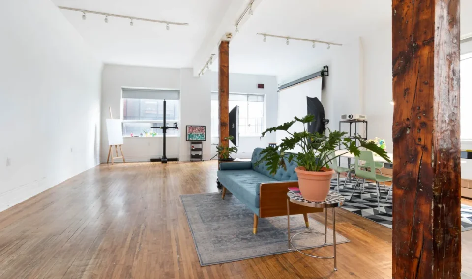 A spacious, well-lit room with wooden floors, large windows, wooden support beams, a blue sofa, potted plants, a rug, and a small table. Art supplies and equipment are visible in the background.