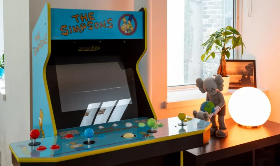 An arcade machine featuring "The Simpsons" stands in a room with a window, a small plant, a soft-lit round lamp, and a figurine on a wooden surface.