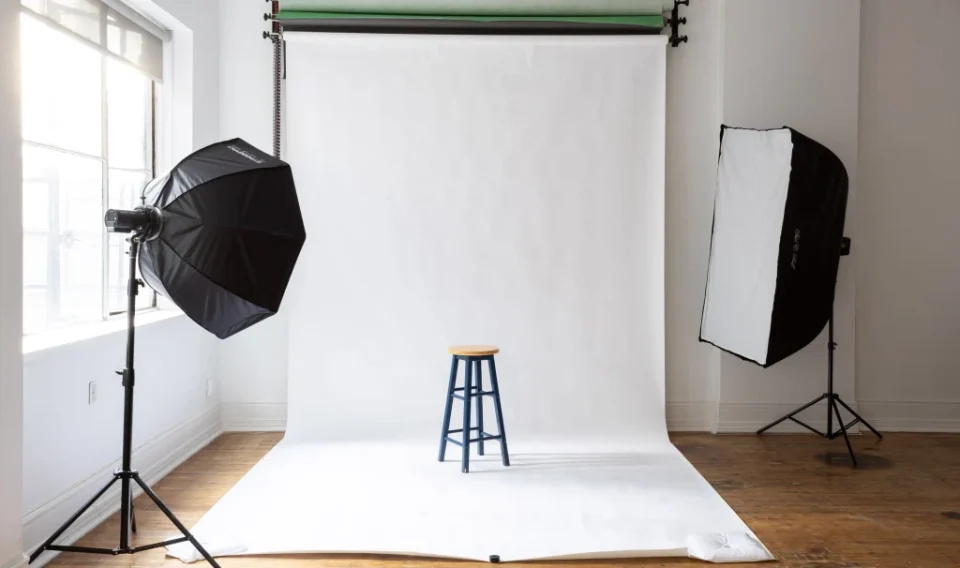 Photography studio setup with a white backdrop, a wooden stool in the center, and two softbox lights on either side.