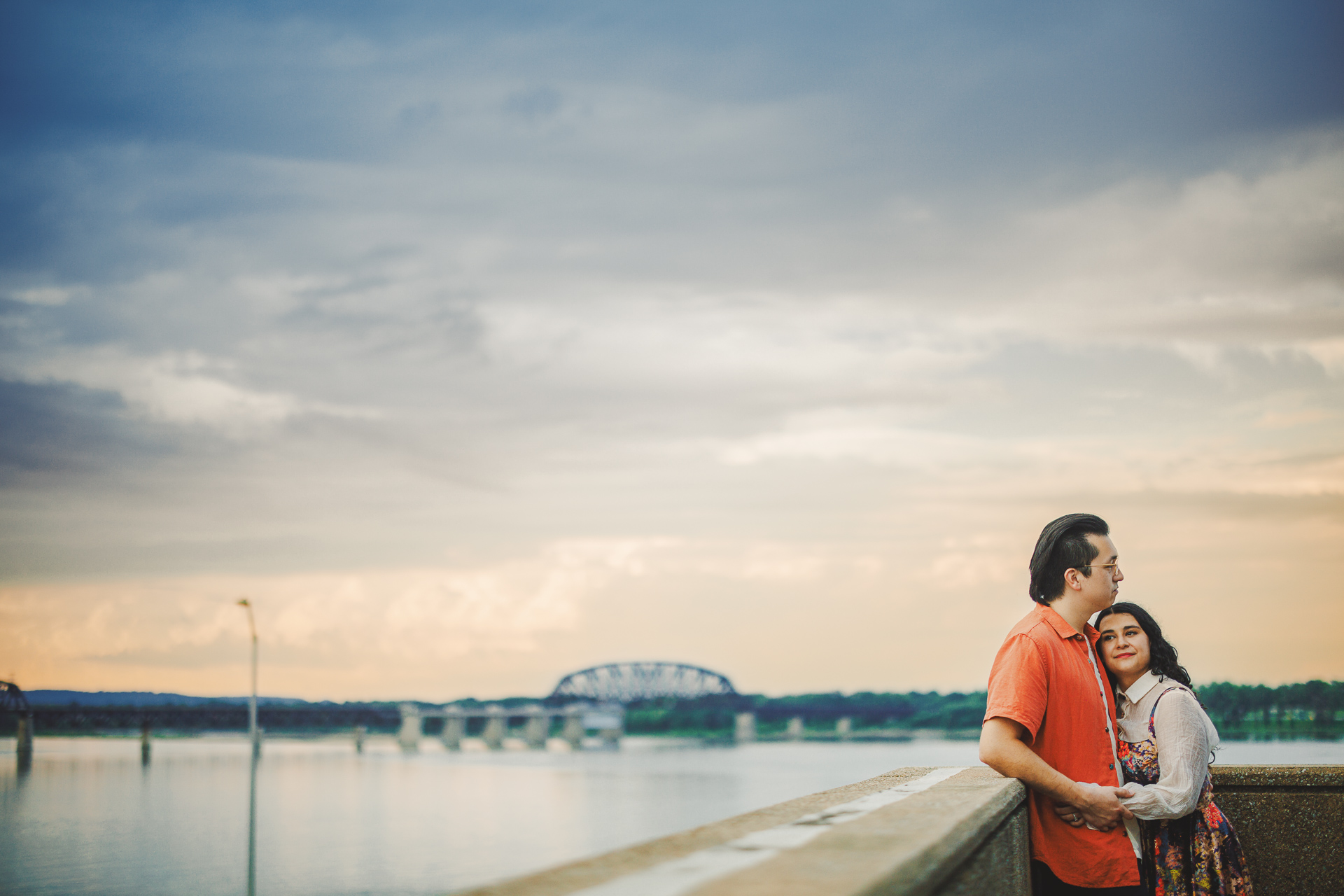 A couple stands on a concrete walkway near the water, with a bridge in the background under a cloudy sky, as if an auto draft of a perfect moment in time.