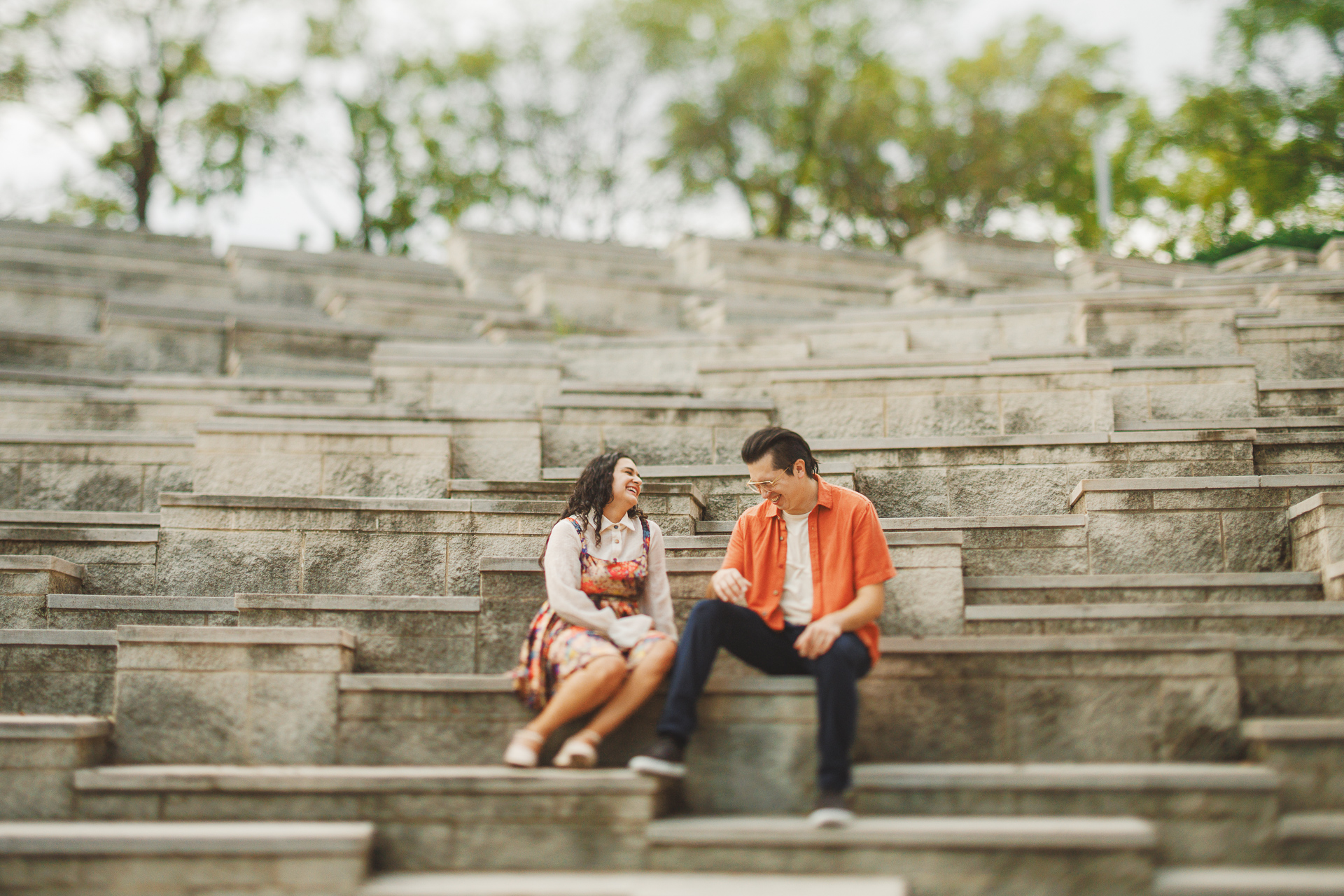 Two people sit and converse on stone steps in an outdoor amphitheater, surrounded by trees. One person is wearing a floral dress, the other is in an orange shirt. With the tranquility of nature around them, their dialogue flows effortlessly, as if scripted by auto draft itself.