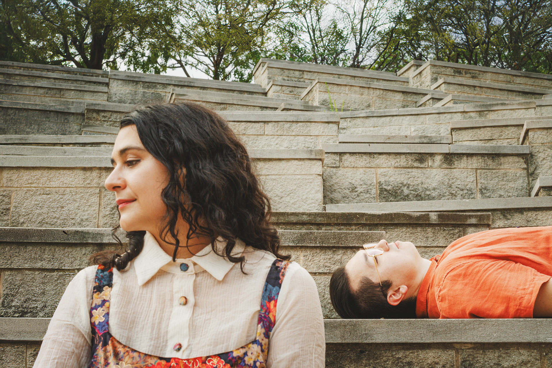 A woman with curly hair sits in the foreground, while in the background, a person in an orange shirt lies on stone steps, as if part of an auto draft scene.