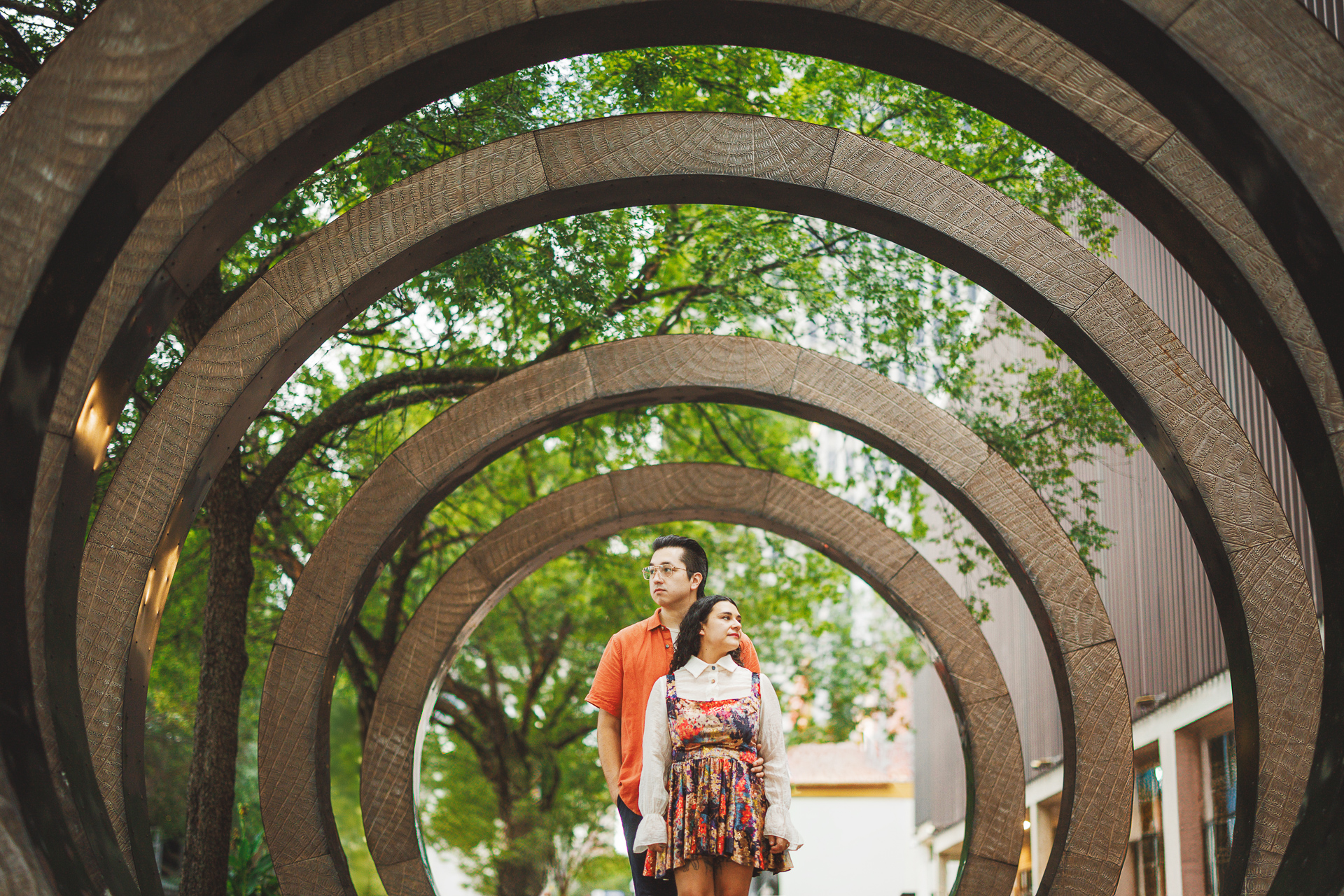 A couple stands underneath a series of large, circular wooden structures in an outdoor park setting, their spontaneity akin to an auto draft selection, with trees gracefully framing the background.