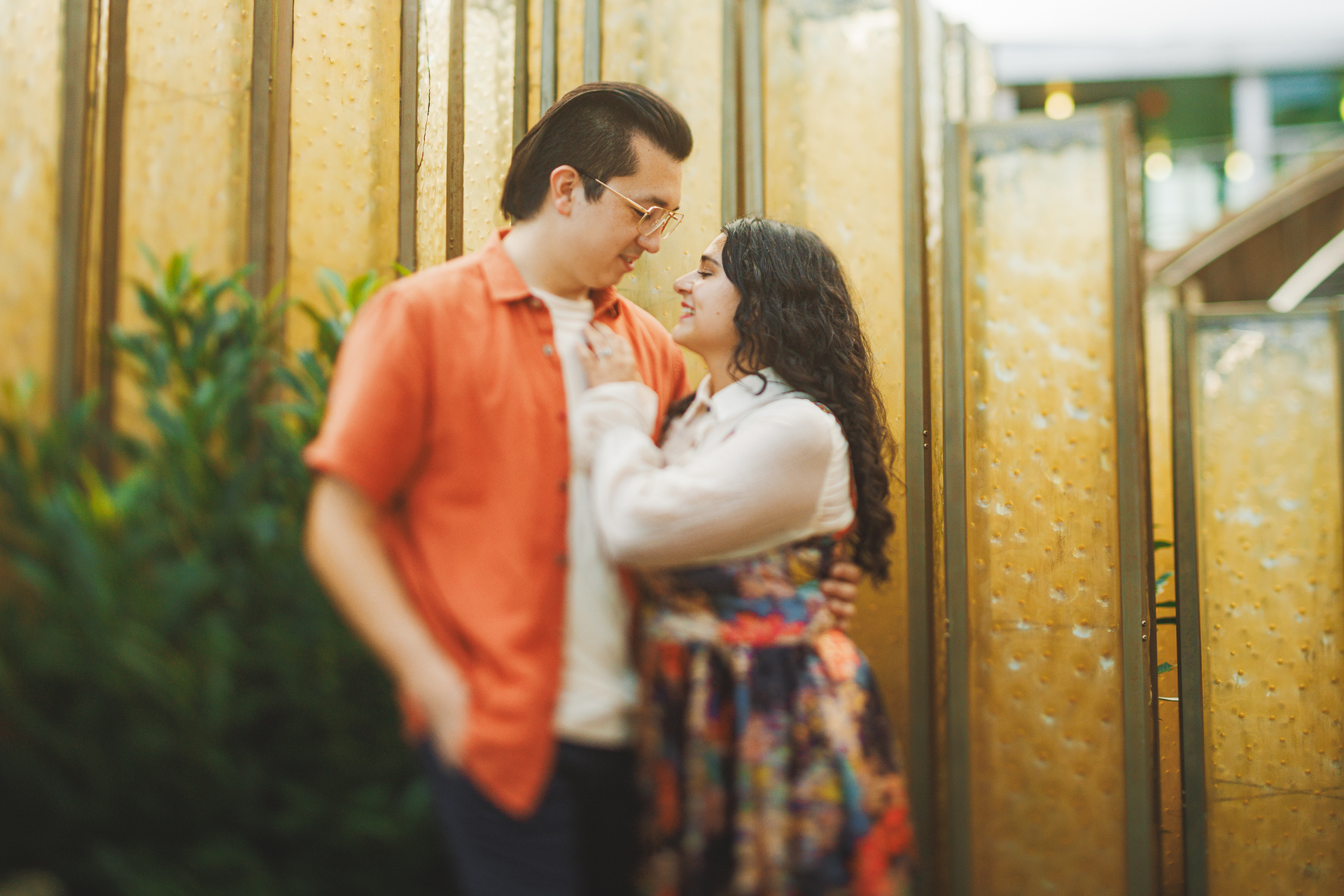 A man and a woman stand close together, smiling and talking. They are indoors near decorative panels and plants. The man, wearing an orange shirt and glasses, seems to draft something while the woman, in a white top with a floral dress, looks on approvingly.
