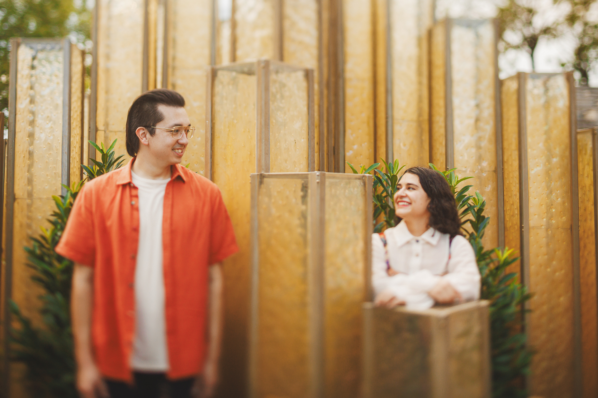 A man in an orange shirt and a woman in a white blouse smile at each other while standing among tall, golden, rectangular structures resembling visions from an Auto Draft sketch.