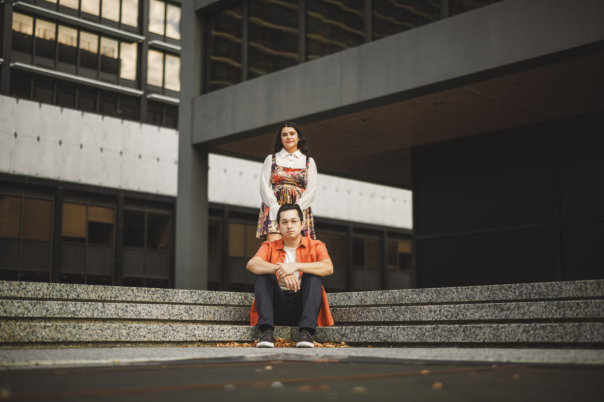 A man in an orange shirt sits on concrete steps, seemingly lost in thought, while a woman in a colorful dress stands behind him. The urban setting with its modern buildings feels meticulously designed, almost like an Auto Draft of contemporary life.