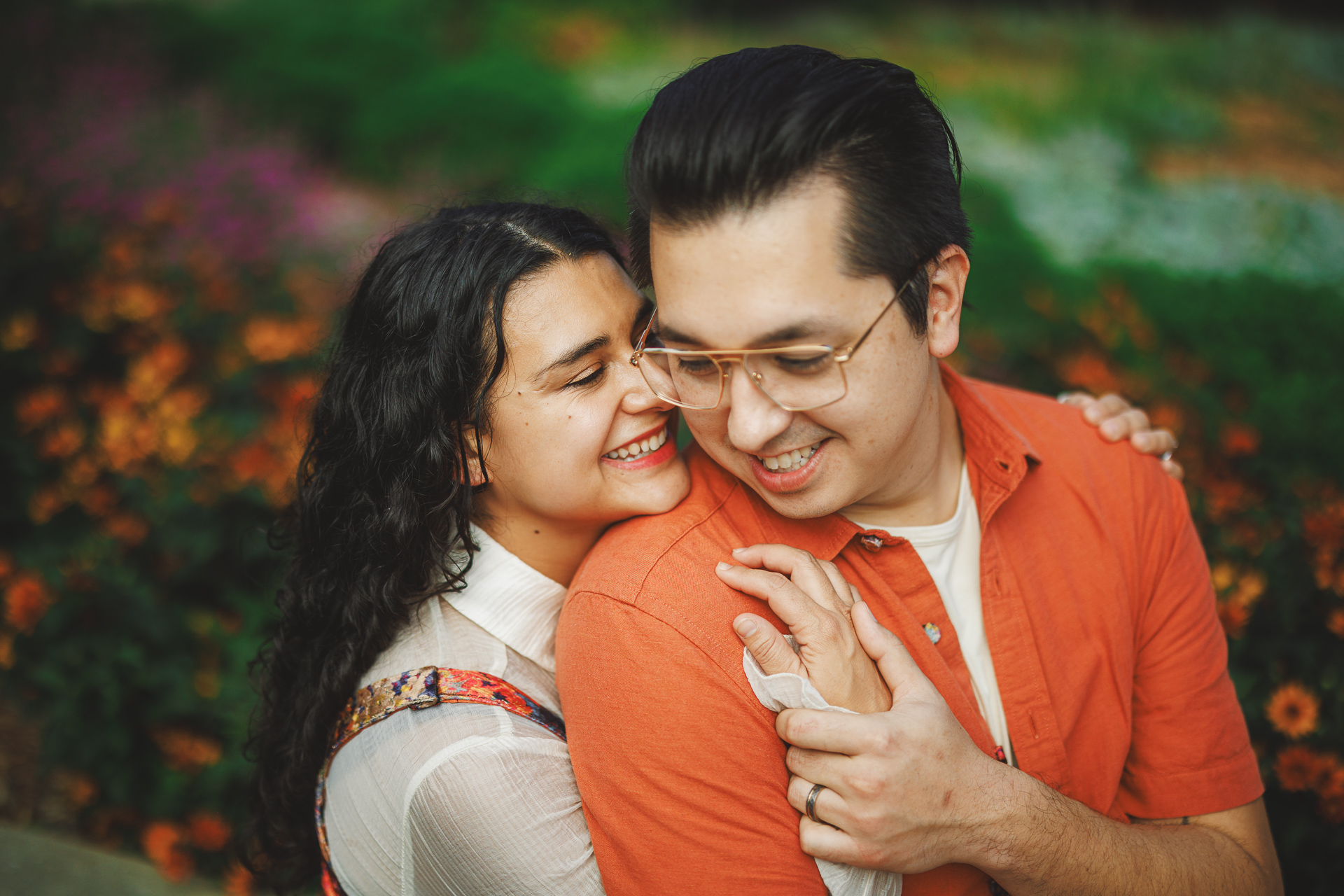 Two people holding each other and smiling outdoors with blurred greenery and flowers in the background, capturing a moment of pure joy.