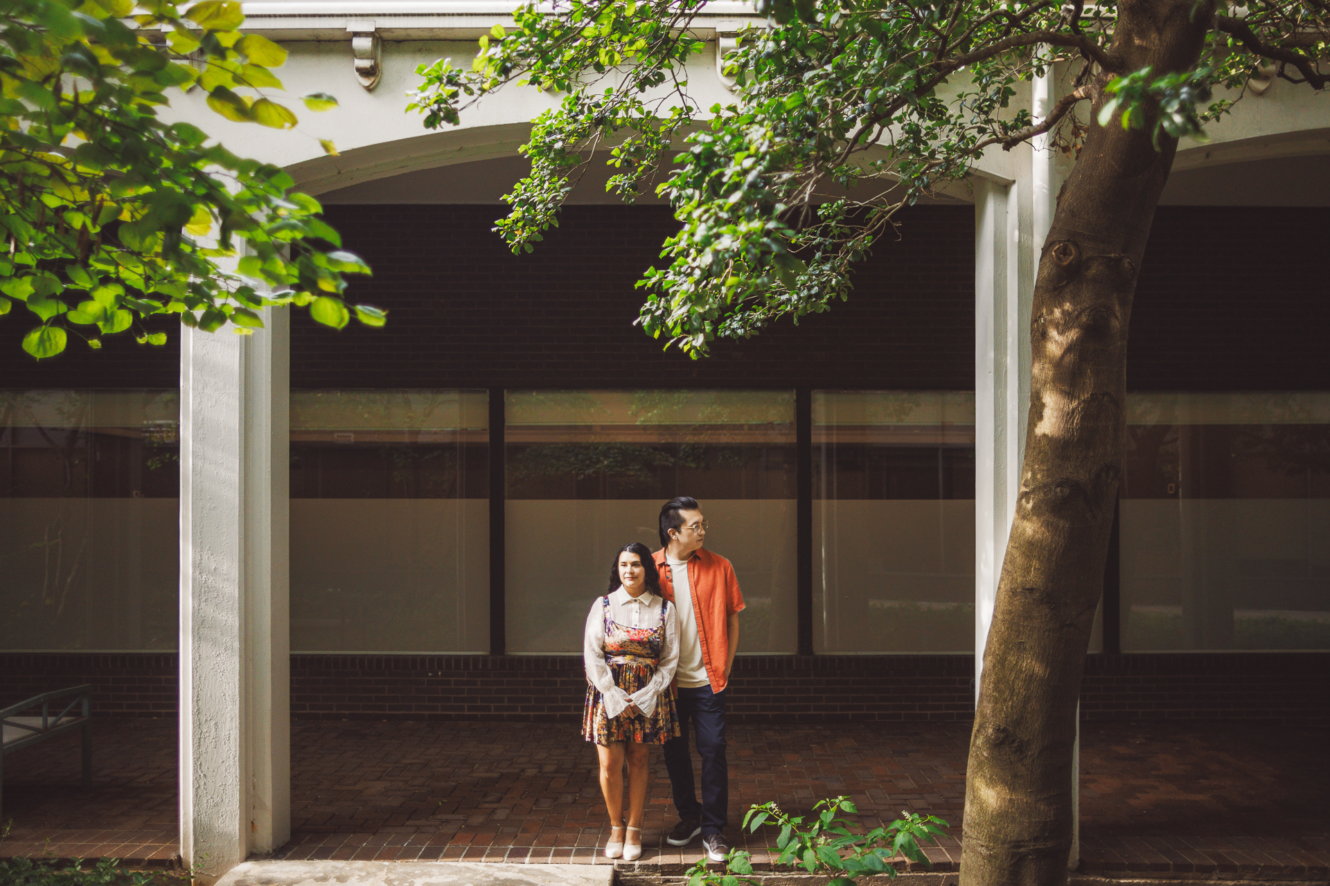 A woman and a man stand side by side under an archway in a courtyard with trees, as if part of an Auto Draft. The man looks at the woman while she gazes forward.