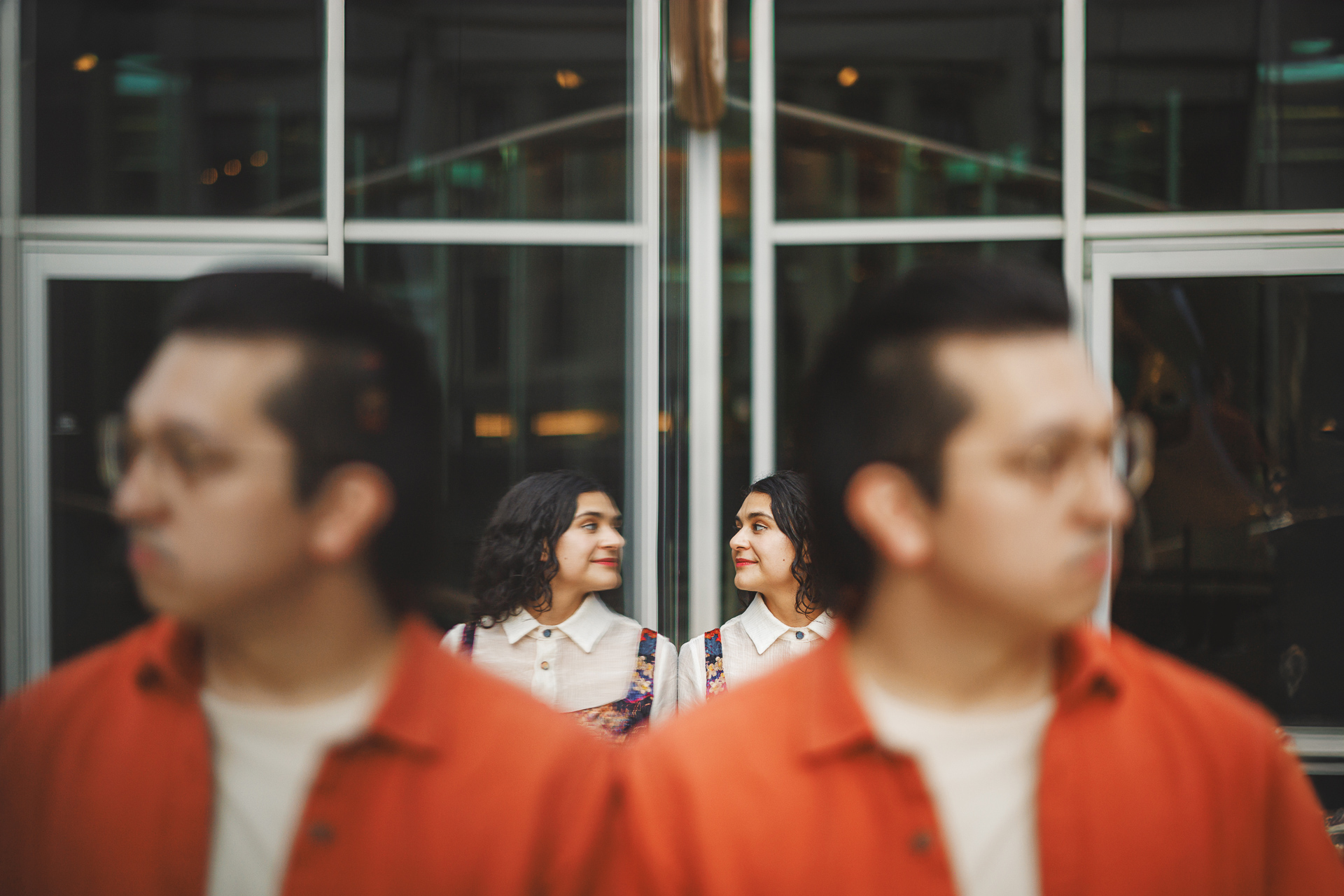 A man in an orange shirt stands with his reflection in the foreground. Two women in white blouses and patterned dresses smile at each other in the background, creating a symmetrical scene that looks like it could be an auto draft of a beautifully composed photograph.