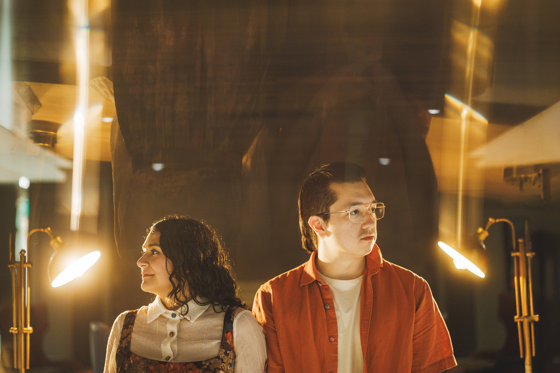Two individuals are standing in a warmly lit room with table lamps, both looking in different directions. One person is wearing a white shirt and floral dress, the other in an orange shirt with glasses, as if contemplating their next move like an auto draft plan.