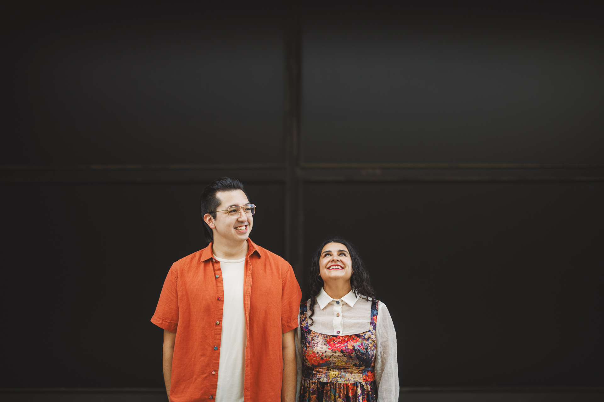 A man and a woman stand together against a dark background. The man, wearing an orange shirt and glasses, seems as if he's contemplating the next Auto Draft. The woman, dressed in a colorful dress with a white collar, beams beside him. Both are smiling and looking up.