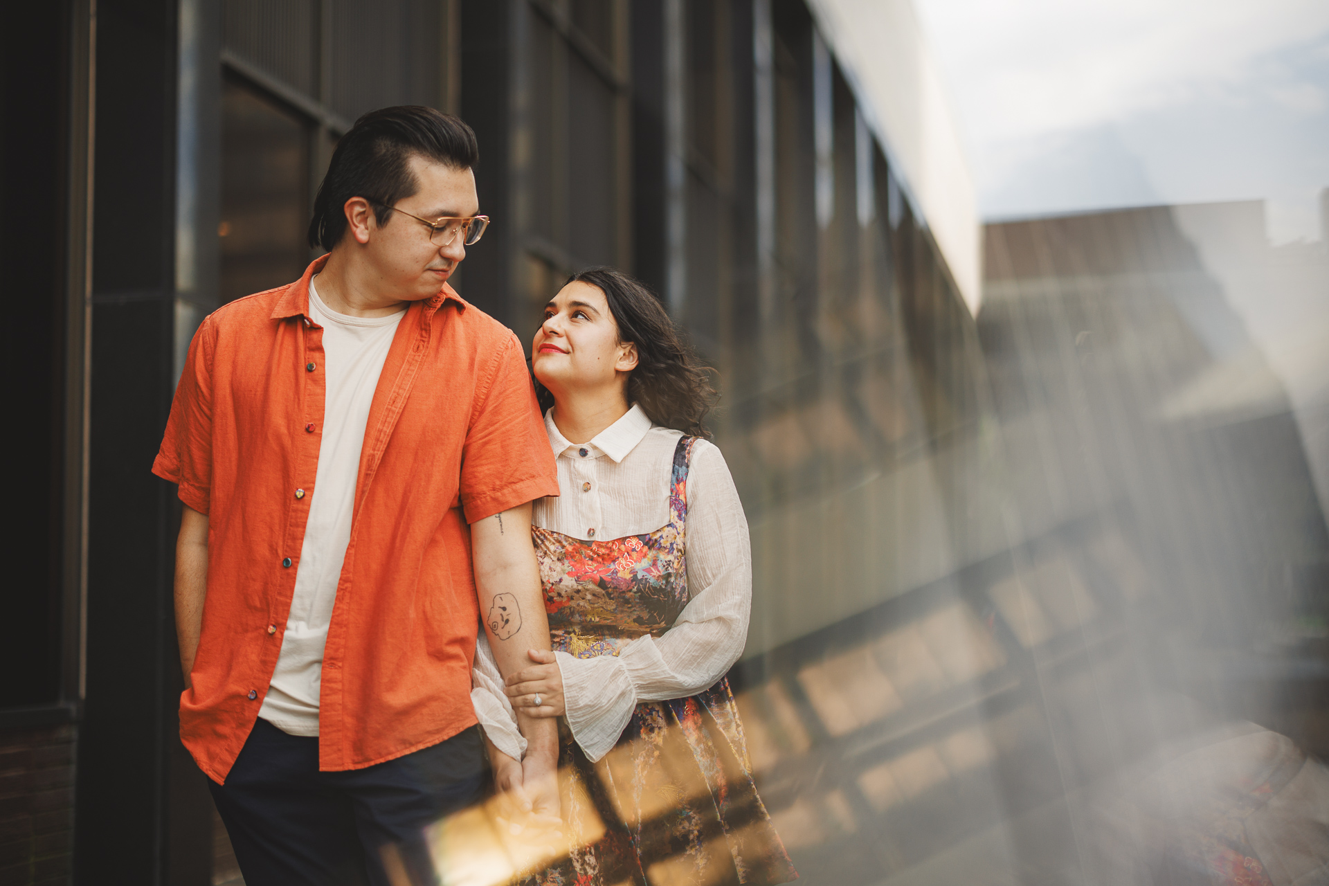 A couple stands close together outdoors. The man wears an orange shirt over a white t-shirt, and the woman, in a flower-patterned dress, looks up at him, holding his arm. In the background of this candid shot captured with Auto Draft precision is a modern building.