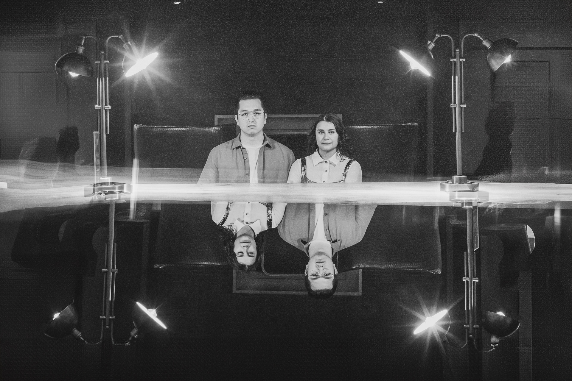 A black and white photograph of two people standing side by side in front of a reflective surface. They are indoors, flanked by four lamps, with their reflections visible below, almost like an artful Auto Draft capturing a moment in time.