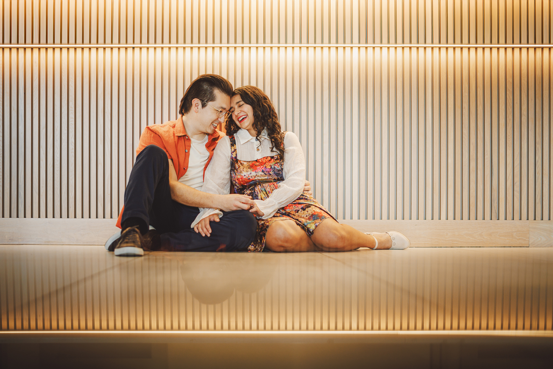 A couple sits on the floor, leaning against each other and smiling, in front of a wooden paneled wall with warm lighting, capturing their love like an auto draft stored fondly in memory.