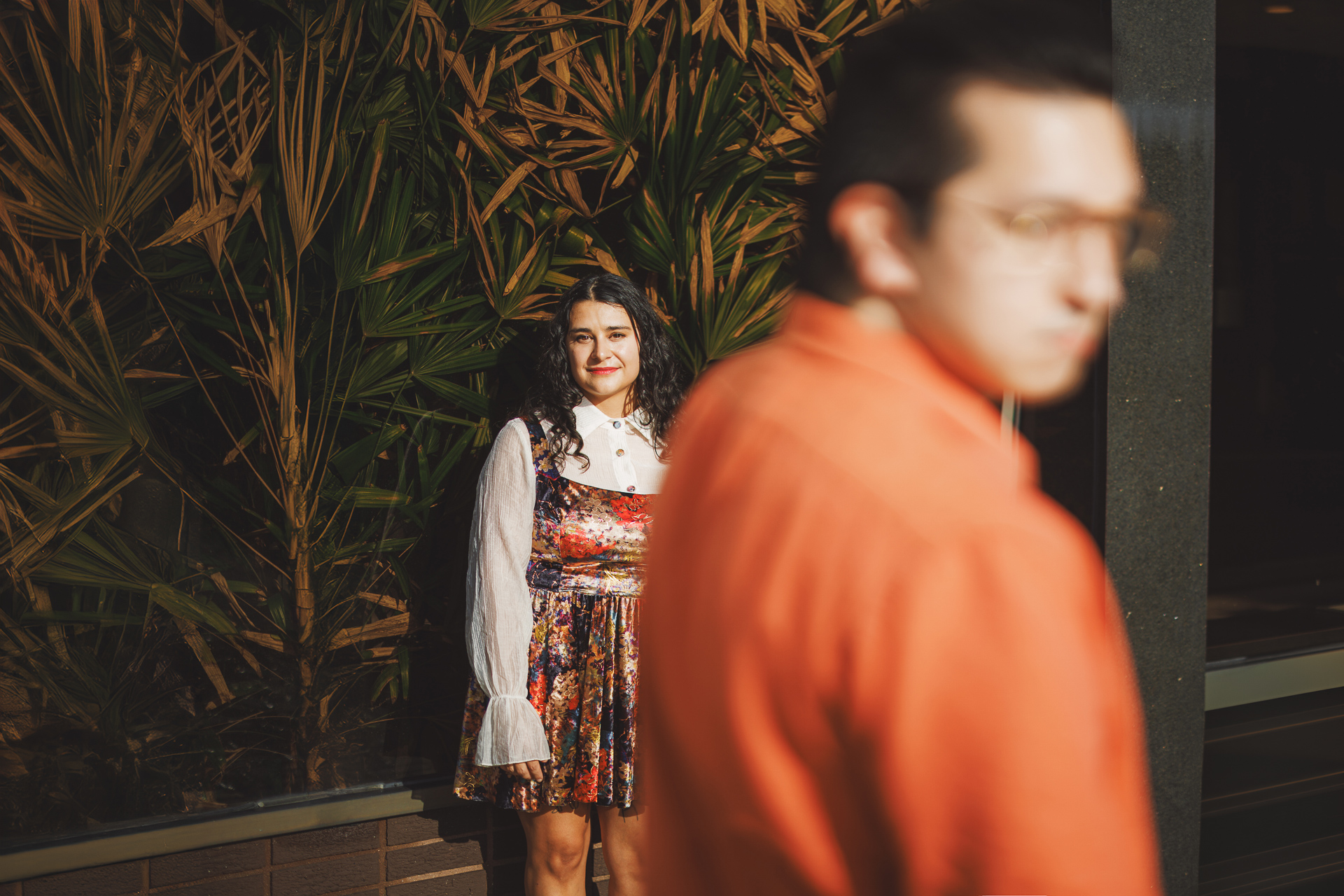 A woman in a colorful dress stands in front of foliage, smiling. In the foreground, a man in an orange shirt is partially visible and slightly out of focus, almost like an auto draft effect.