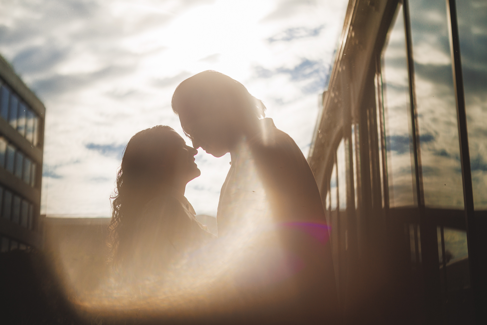 Two people standing close, facing each other, silhouetted against a bright, partly cloudy sky with reflections in nearby glass windows create a scene that feels like an auto draft of a moment suspended in time.